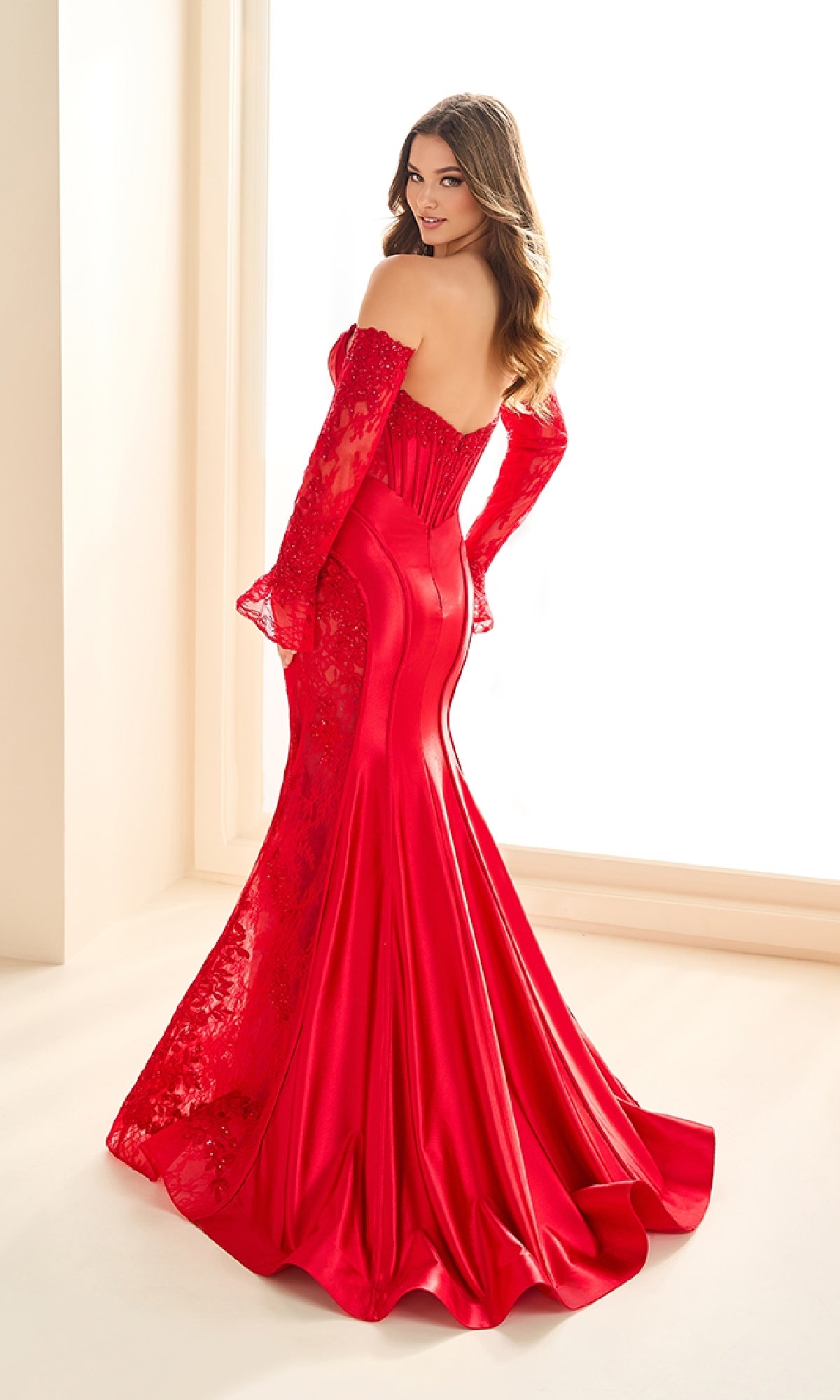 Woman in a fitted, off-the-shoulder, floor-length red Ellie Wilde EW36010 prom dress with lace sleeves and panels stands before a bright window, looking over her shoulder.